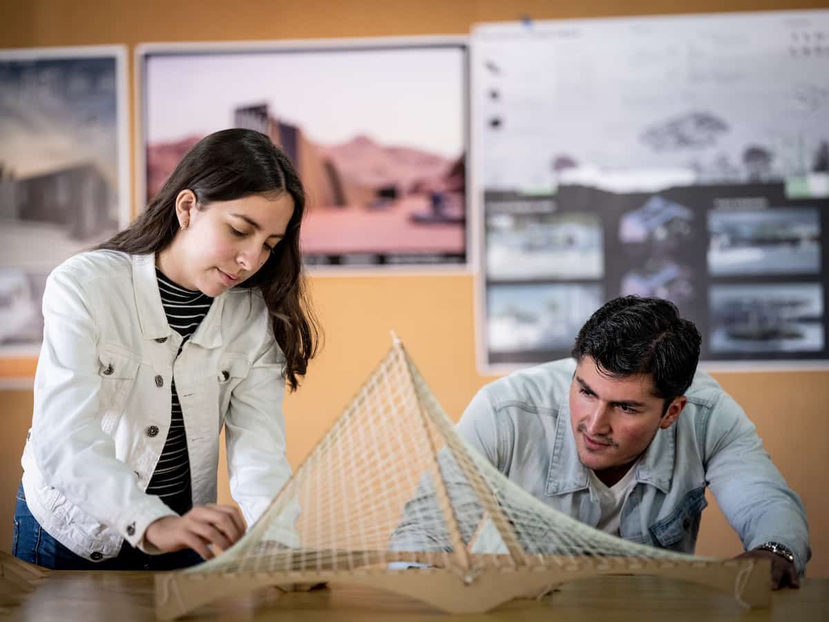 Estudiantes de arquitectura ajustando maqueta estructural con tensores de hilo sobre mesa de trabajo.