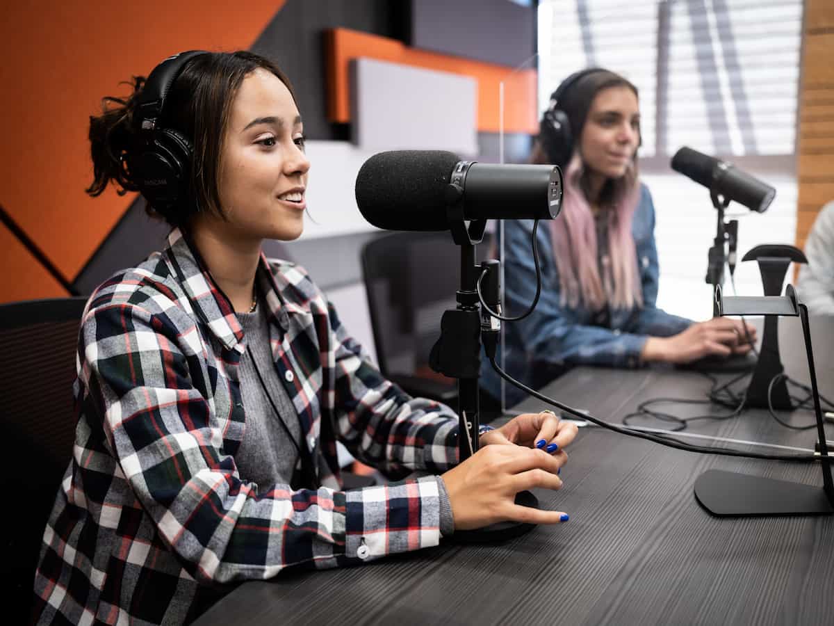 Joven hablando frente a micrófono mientras graba podcast junto a compañera en estudio con paneles acústicos.