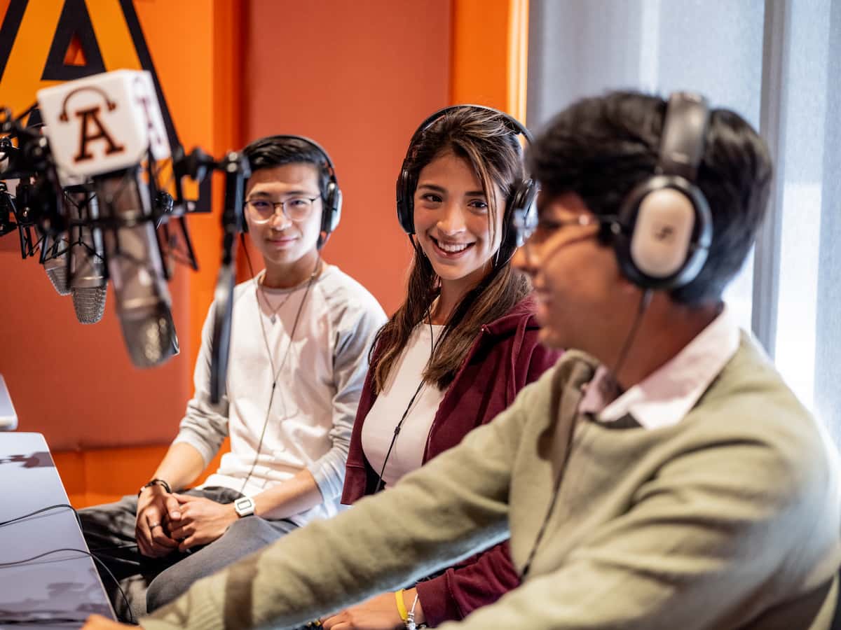 Estudiantes de comunicación grabando contenido radial en cabina profesional con logo institucional al fondo.