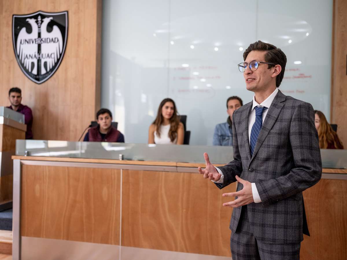 Estudiante de Derecho exponiendo un caso en un aula tipo tribunal, frente a compañeros que simulan un jurado, representando práctica jurídica, argumentación oral y formación legal profesional.