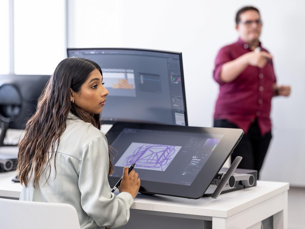 Estudiante concentrada en su trabajo de diseño digital usando una tableta gráfica, mientras el profesor imparte una clase en el fondo del aula.