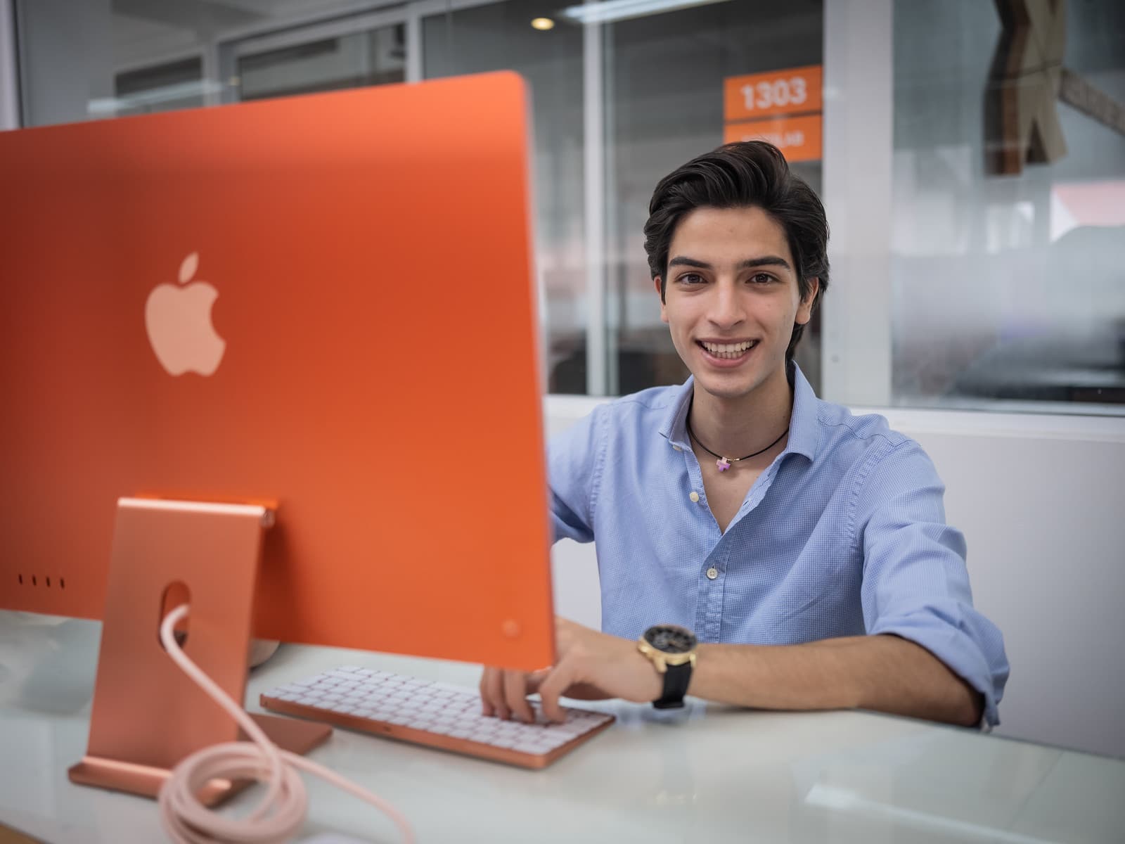 Estudiante sonriendo frente a una computadora de escritorio naranja en un aula acristalada, usando teclado y mouse.