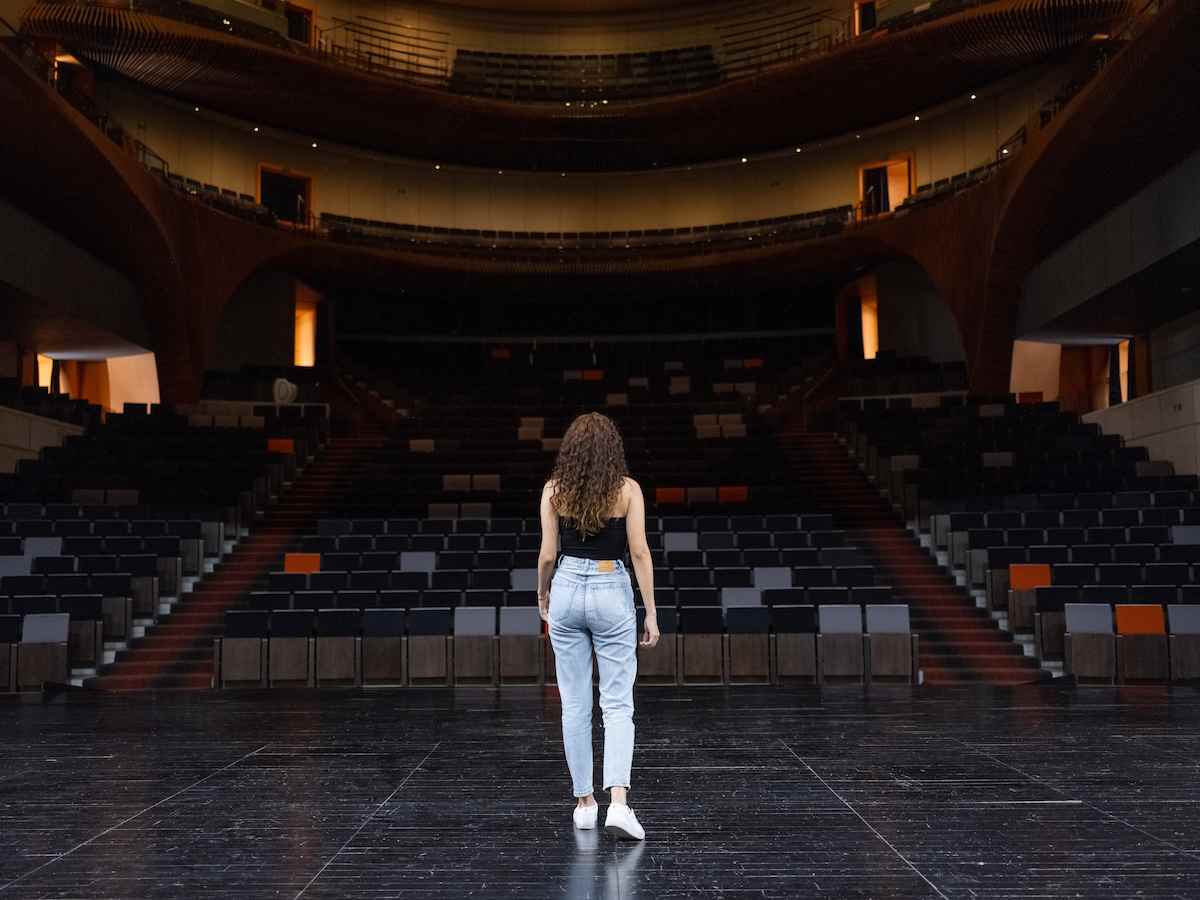 Mujer de espaldas con cabello rizado, camiseta negra y jeans claros, de pie en el centro de un amplio escenario de teatro moderno, observando la sala vacía de butacas divididas en varios niveles, en un momento de contemplación o preparación.