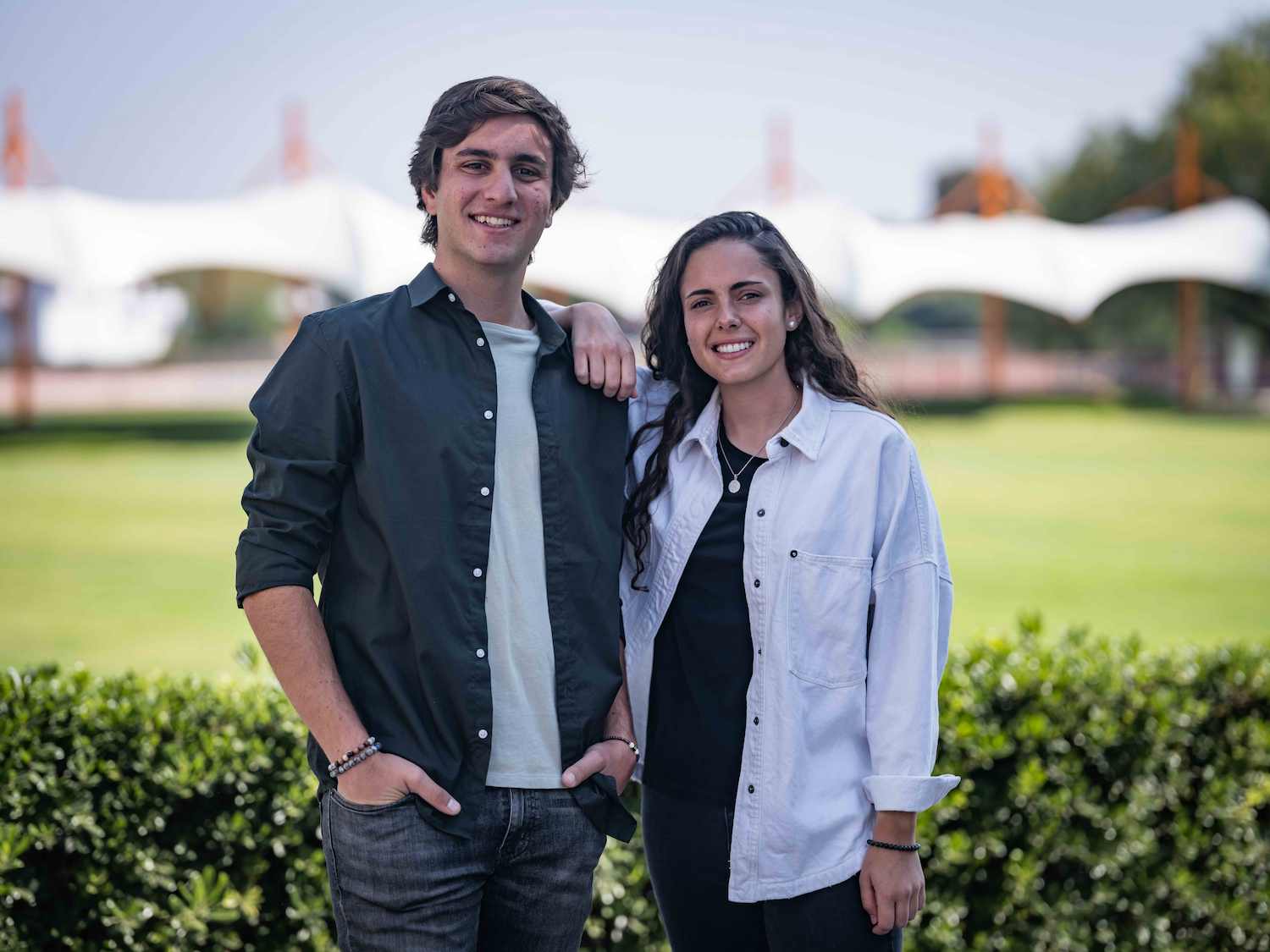 Dos estudiantes sonrientes posan al aire libre frente a una estructura blanca tipo toldo en un campus universitario, con áreas verdes al fondo.