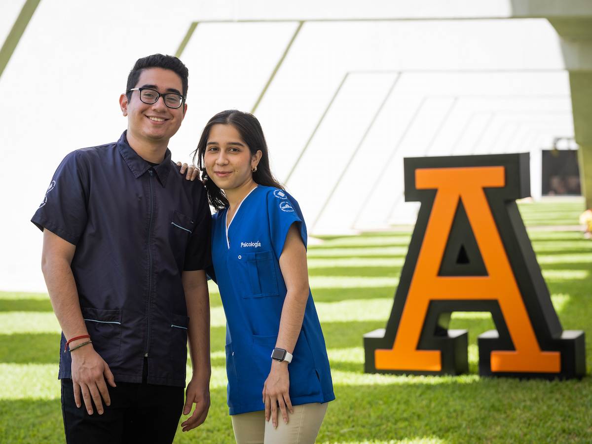 Estudiantes de Psicología posando sonrientes con uniforme clínico frente al logotipo universitario, reflejando vocación de servicio, formación profesional y compromiso con el bienestar emocional.