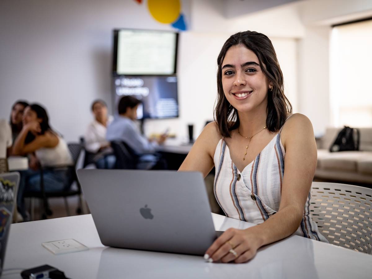 Estudiante de Psicopedagogía sonriendo mientras trabaja en su laptop en un aula colaborativa, reflejando formación integral, acompañamiento educativo y uso de herramientas digitales en el aprendizaje.
